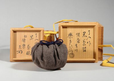 Eine schwarze Teeschale aus Raku-Keramik von Kichizaemon IX. Hier in seinem Stoffbeutel mit den dazugehörigen Holzdosen und deren Beschriftung.